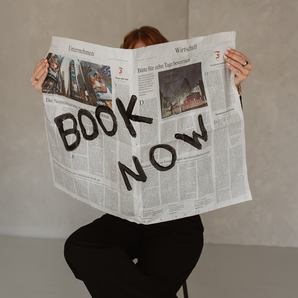 Zwei Hände halten eine Zeitschrift mit der Aufschrift Book Now.